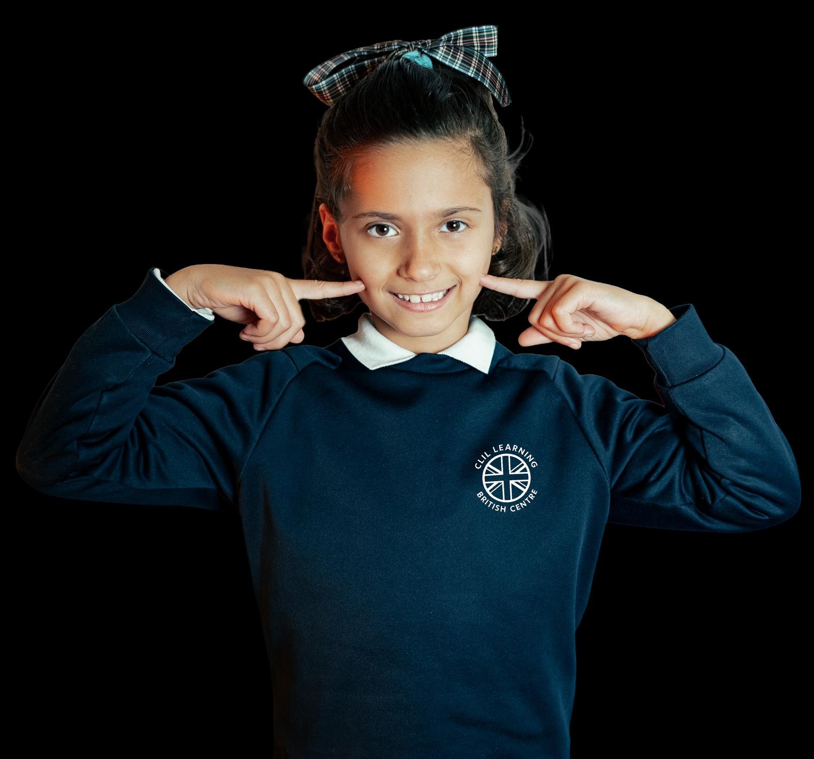 Niña sonriendo y señalando mejillas en uniforme escolar.